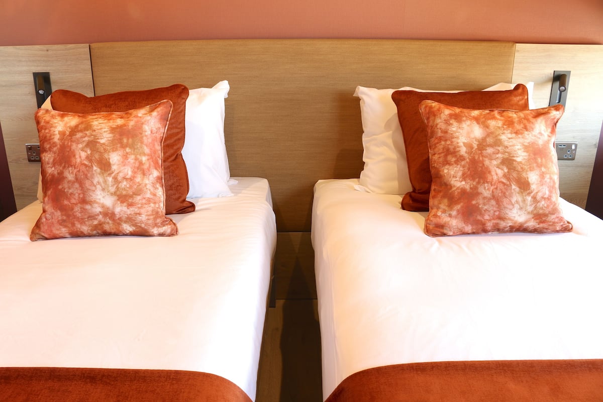 Two twin beds with matching brown and white bedding, a wooden headboard, and a control panel on the wall.