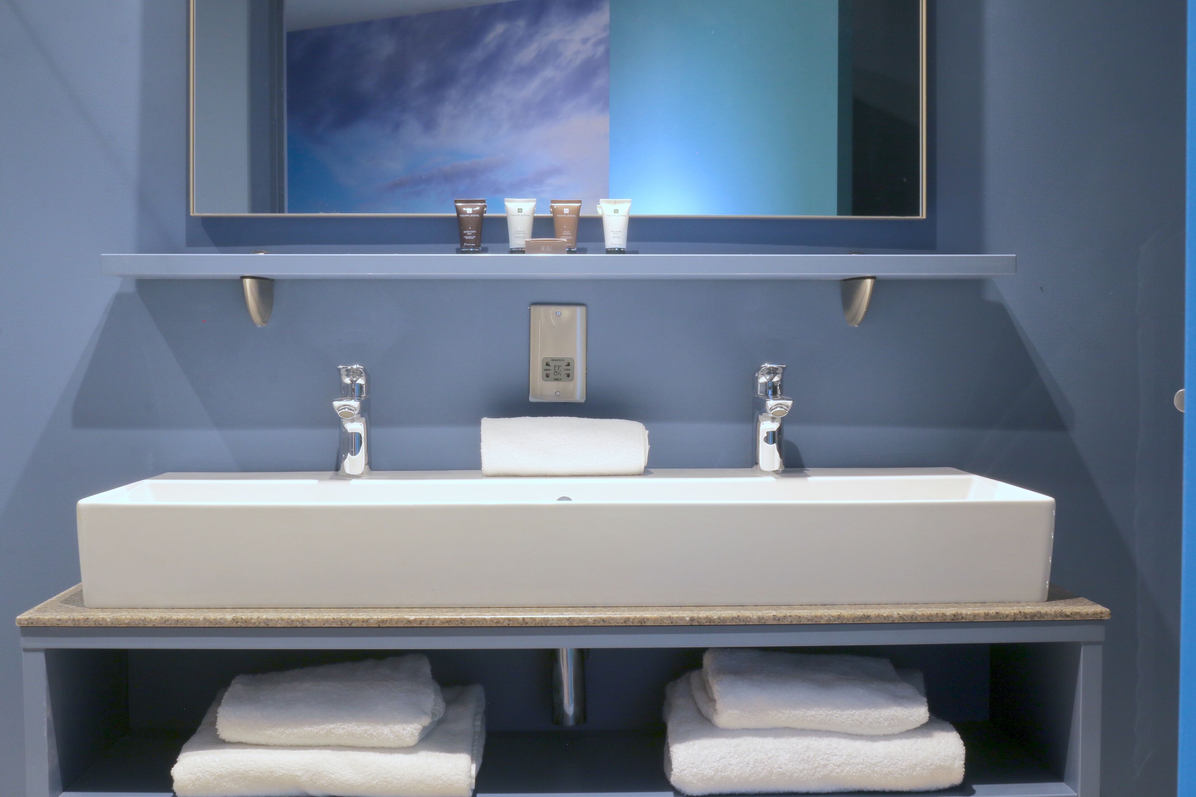 A modern bathroom with a large sink, two faucets, and a mirror above. There are three toiletries on the sink's edge and a towel rack below.