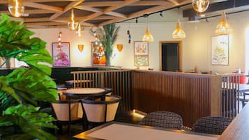 A restaurant with wooden ceiling beams, hanging pendant lights, and a central wooden counter. There are round tables with wicker chairs, and potted plants are visible.