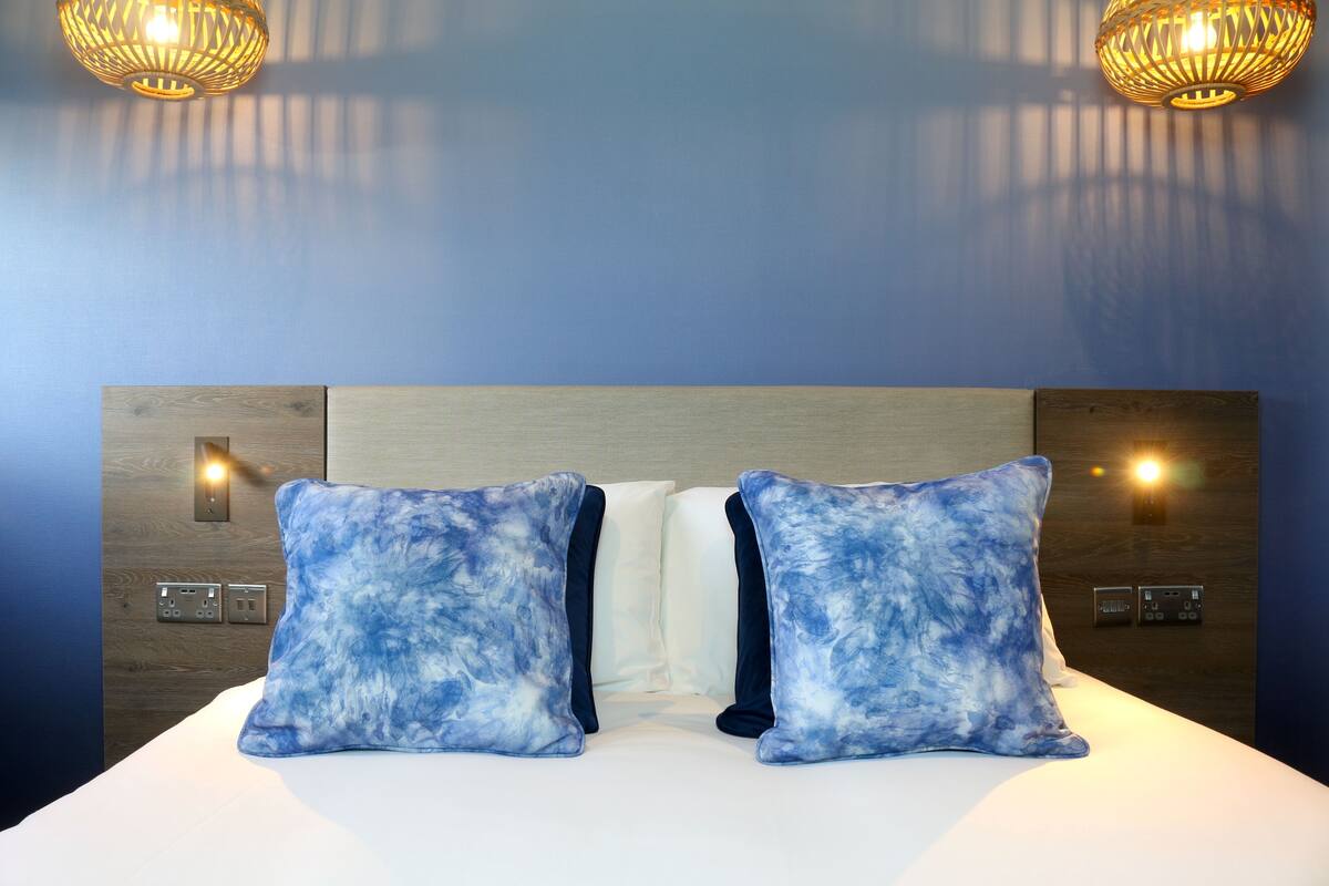 A hotel room with a bed, two blue tie-dye pillows, a wooden headboard, wall-mounted light fixtures, and electrical outlets.