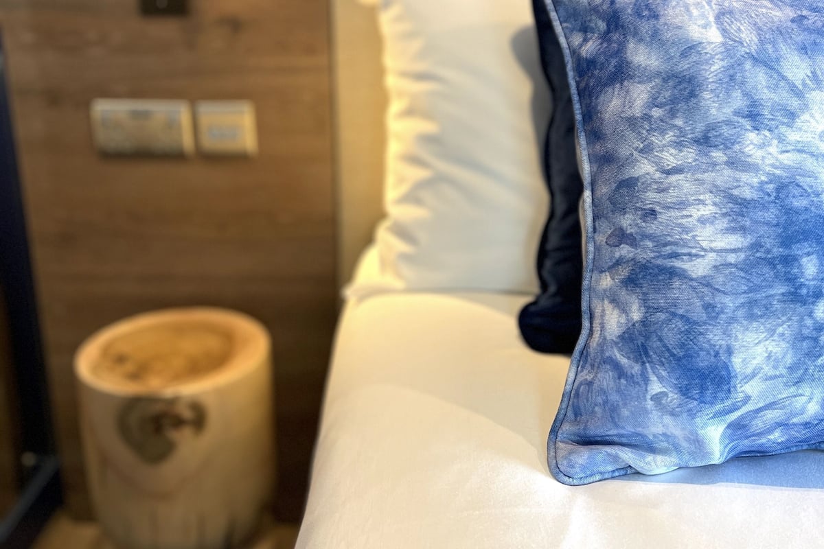 A bed with white bedding and a blue patterned pillow, a wooden nightstand with a lamp, and a wall-mounted light fixture.