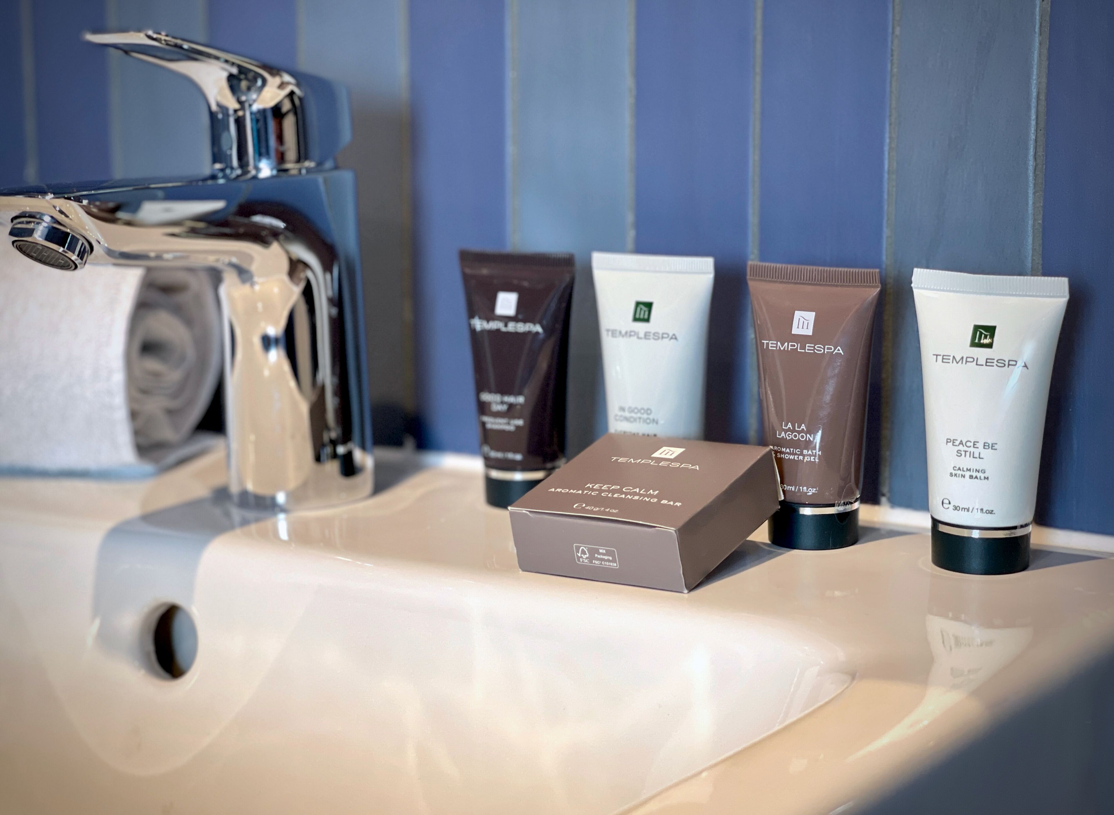 A bathroom sink with toiletries including a cleansing bar, a shower gel, and a calming skin balm.