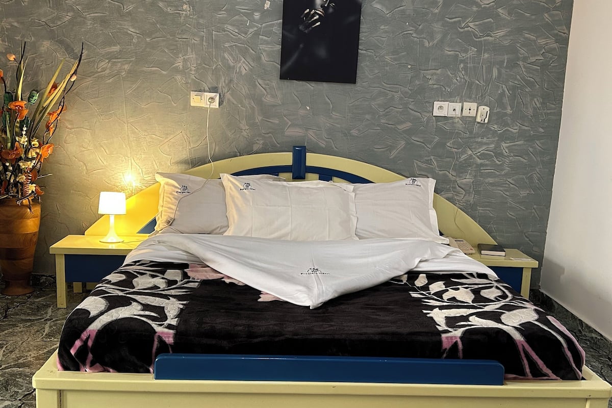A bed with a blue frame, white pillows, and a black and white patterned blanket. There is a nightstand with a lamp, a wall-mounted power outlet, and a framed picture.