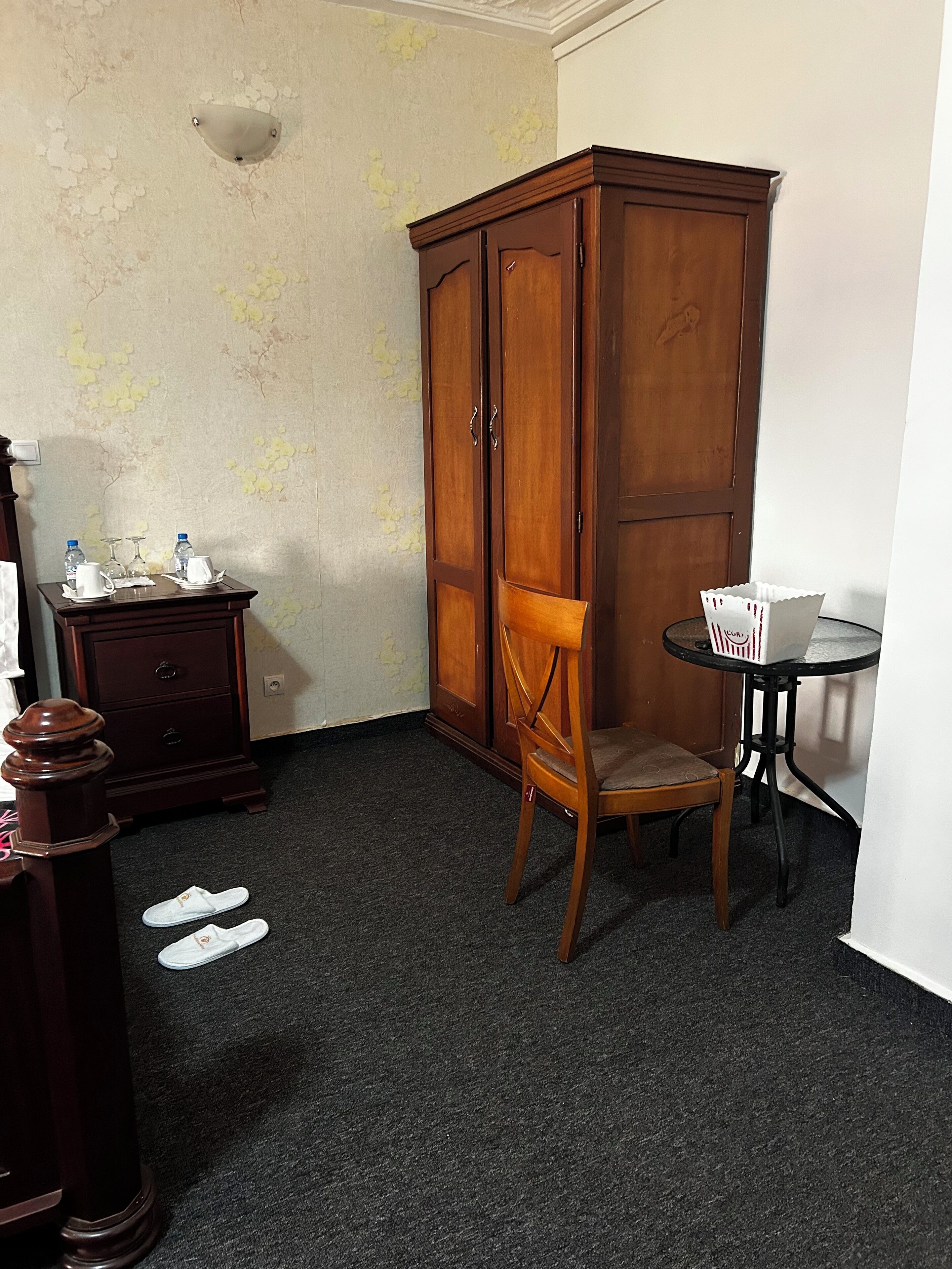 Una habitación de esquina con un armario de madera, una mesita con una cesta, una silla y un par de zapatillas en el suelo.