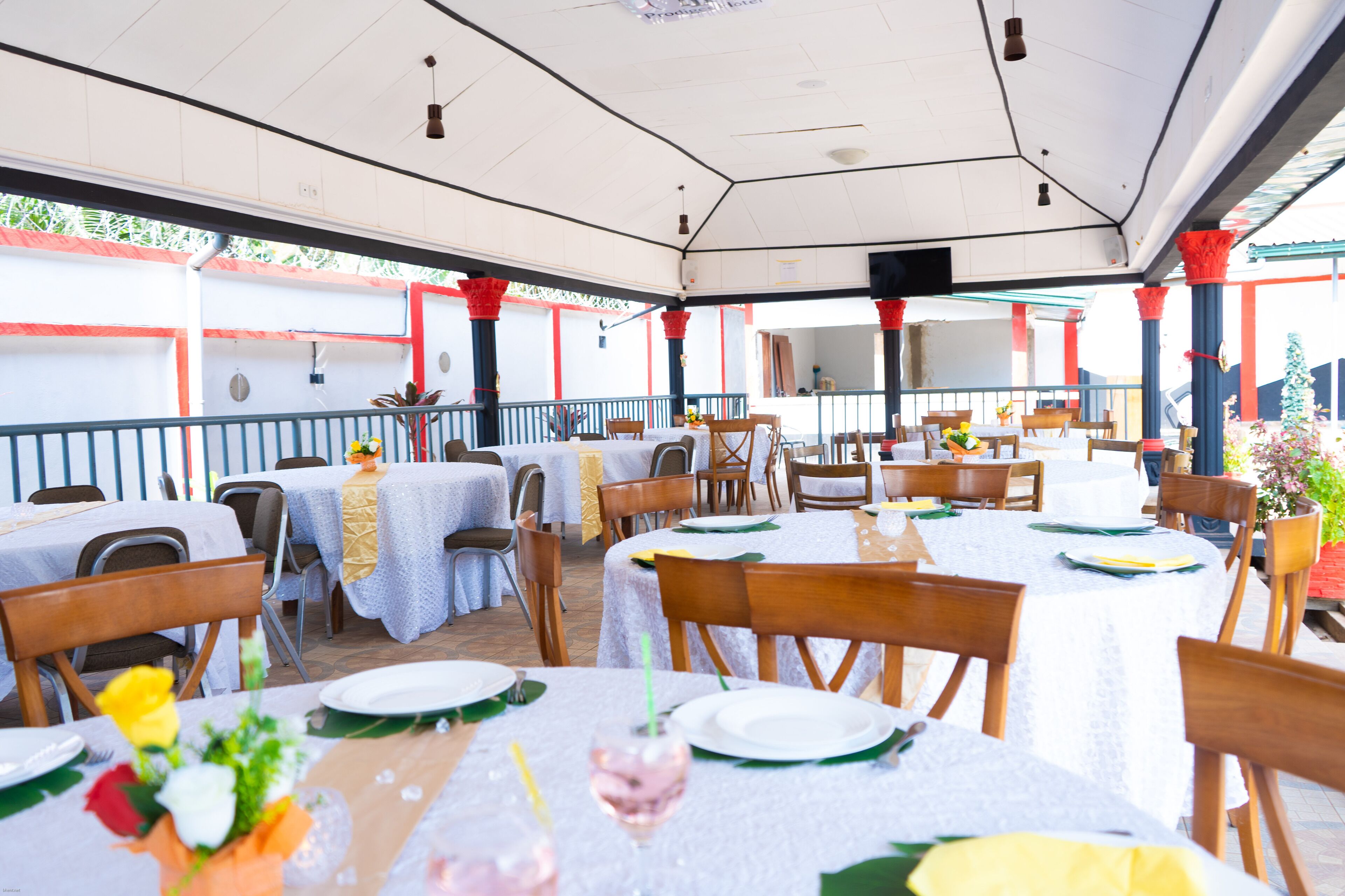 Un espacio de comedor al aire libre con mesas dispuestas para comidas, que cuenta con techo cubierto y pilares rojos.