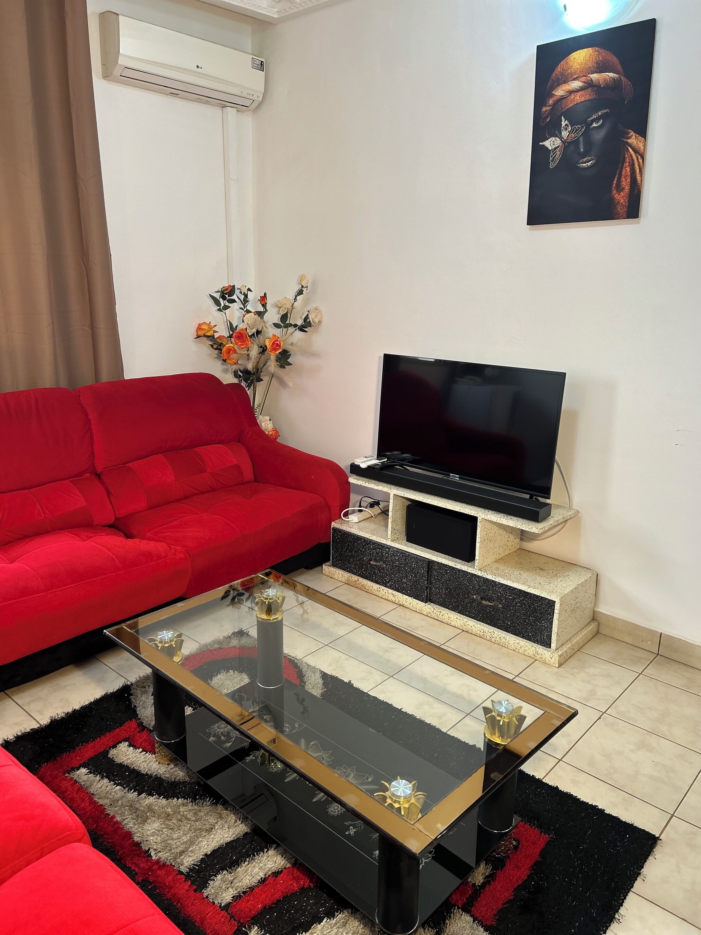 A living room with a red sofa, a glass coffee table, a TV on a stone platform, and a wall-mounted air conditioner.