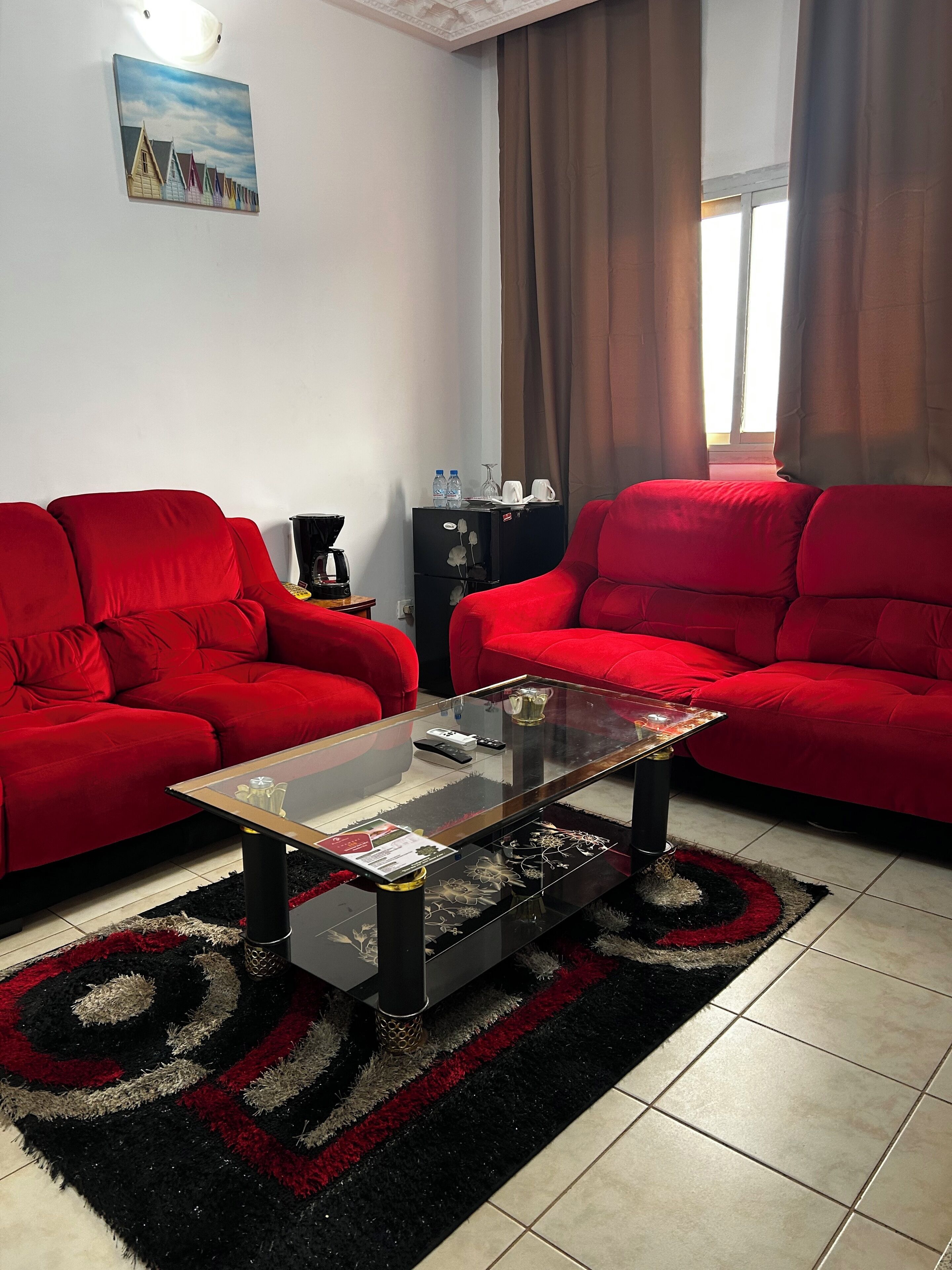 Una sala de estar con dos sofás rojos, una mesa de centro de vidrio, una mesita auxiliar negra con una cafetera y un cuadro en la pared.