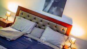 Een net opgemaakt bed met patroonkussens en een geborduurde hoofdbord, twee bedlampjes en een lijst met een kunstwerk aan de muur.