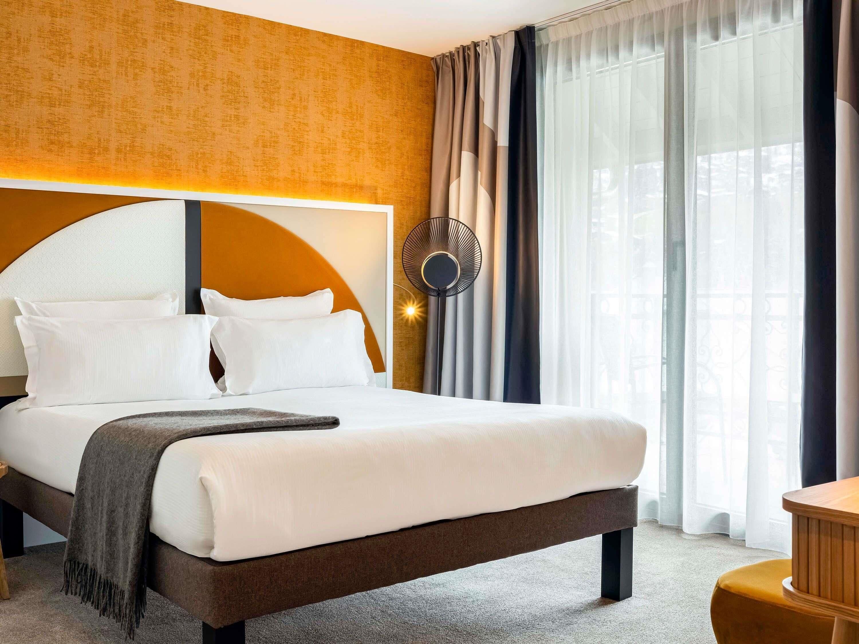 Ein Hotelzimmer mit einem großen Bett, einer runden Wanduhr und Blick ins Freie durch transparente Vorhänge.