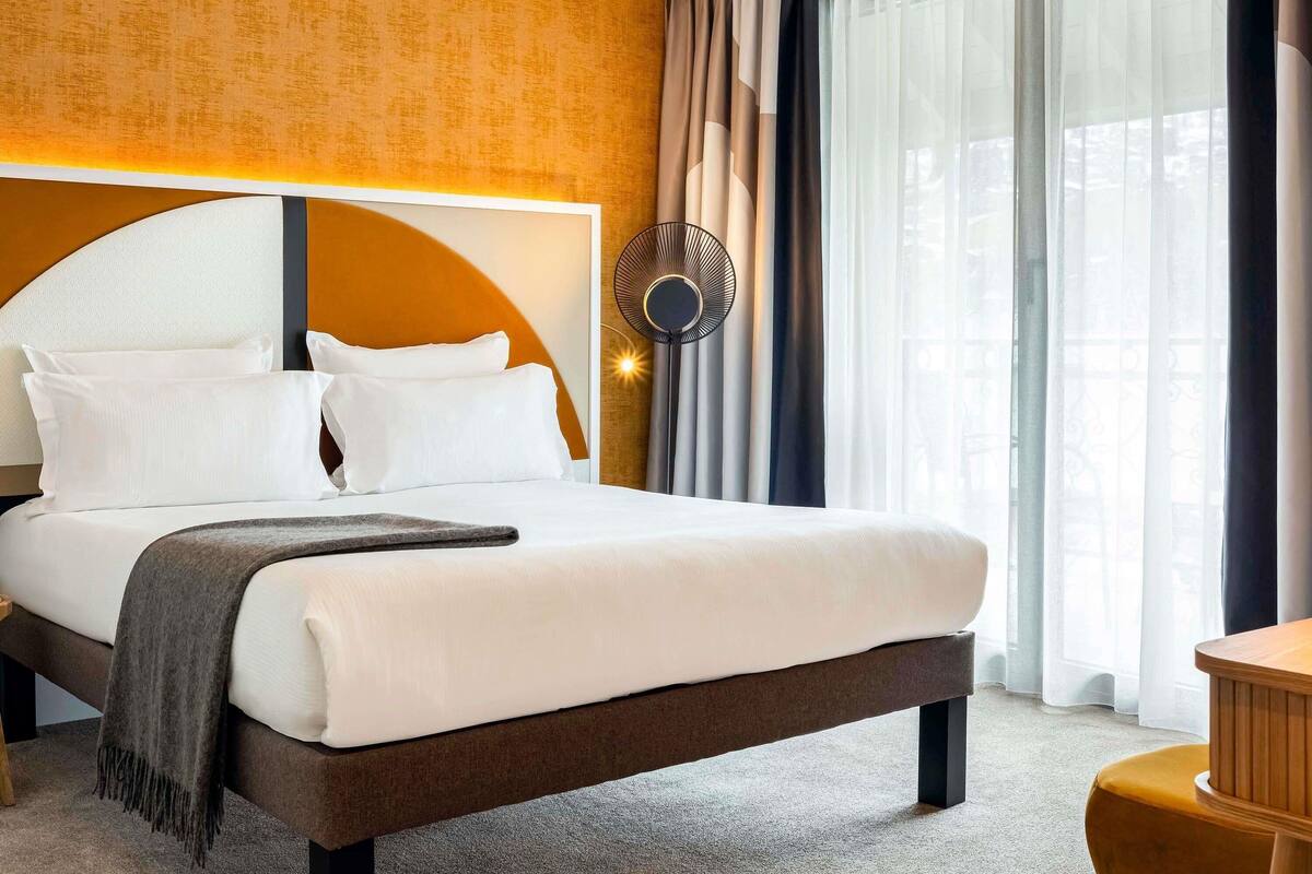 A hotel room with a large bed, a round wall clock, and a view of the outdoors through sheer curtains.