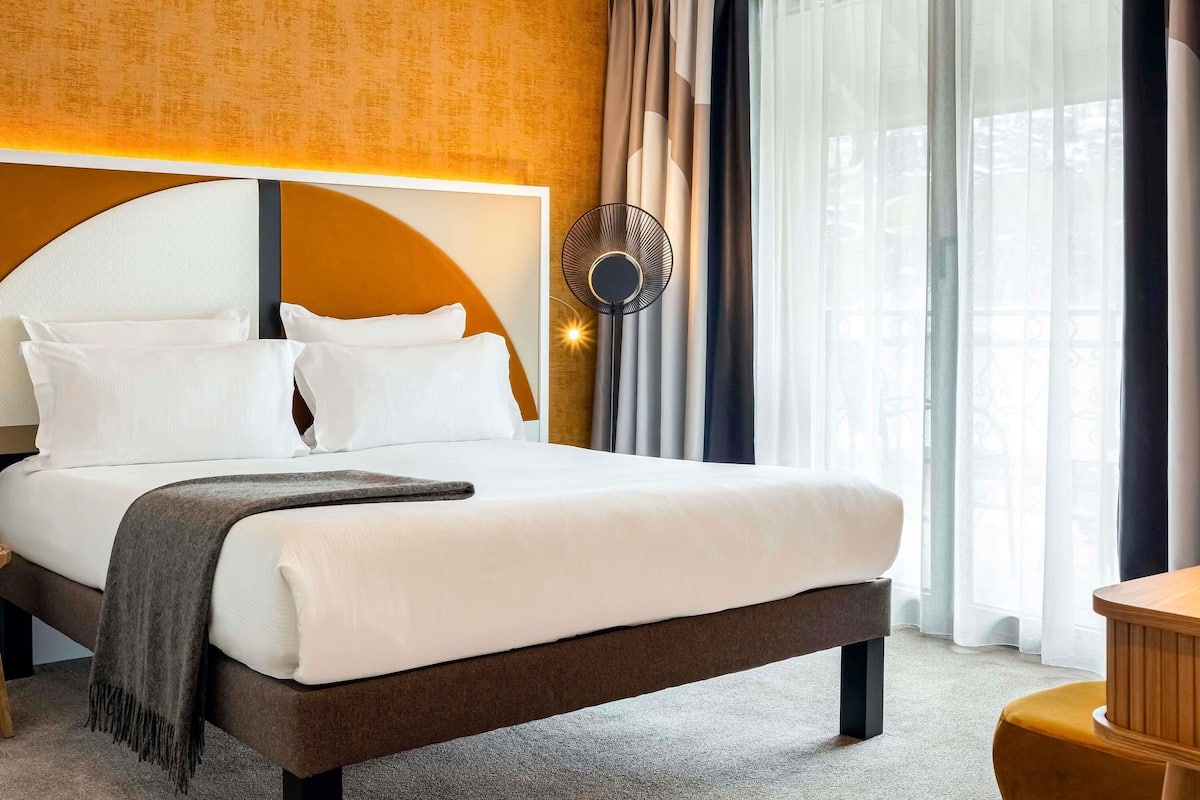 A hotel room with a large bed, a round wall clock, and a view of the outdoors through sheer curtains.