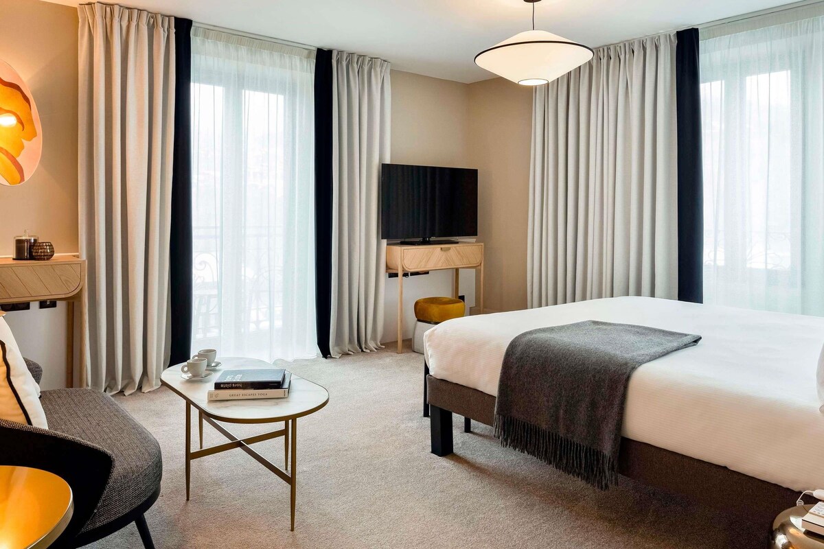 A hotel room with a bed, a desk with a TV, a chair, a small table, and curtains.