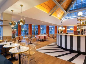 A modern restaurant with round tables, wicker chairs, a central bar, and large windows. - Saint-Gervais Hotel and Spa (Saint-Gervais-les-Bains)