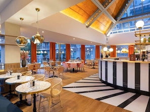 Un restaurant moderne avec des tables rondes, des chaises en osier, un bar central et de grandes fenêtres.