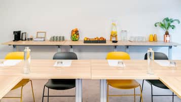 A conference room with a long table, chairs, a coffee machine, fruit basket, and a potted plant.