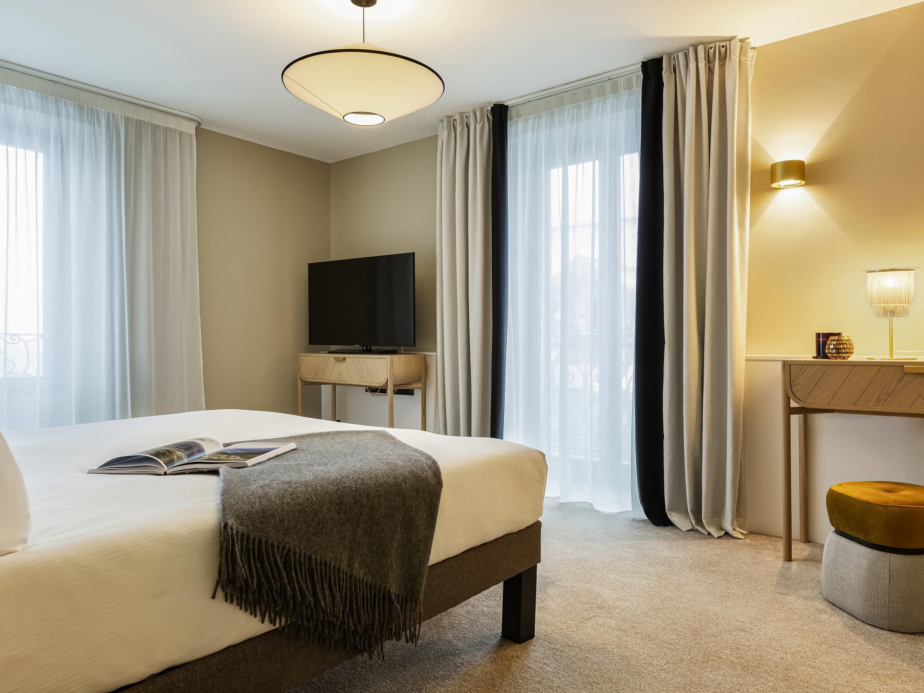 A hotel room with a bed, a flat-screen TV on a wooden stand, a bedside table with a lamp, and a view through sheer curtains.