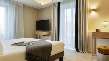 A hotel room with a bed, a flat-screen TV on a wooden stand, a bedside table with a lamp, and a view through sheer curtains.