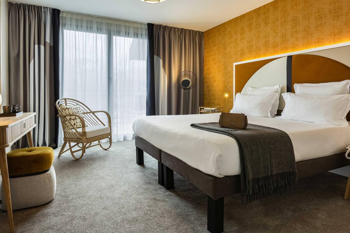 A hotel room with a bed, bedside table, chair, and a view of the outdoors.