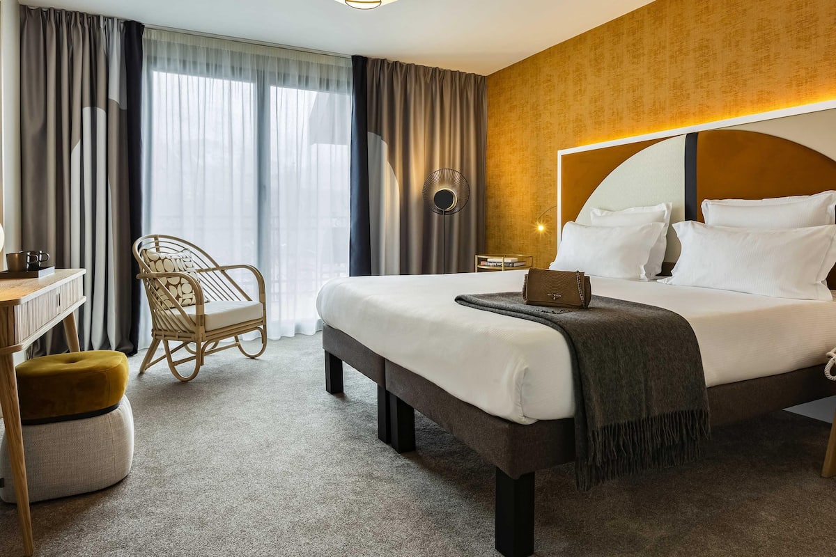A hotel room with a bed, bedside table, chair, and a view of the outdoors.