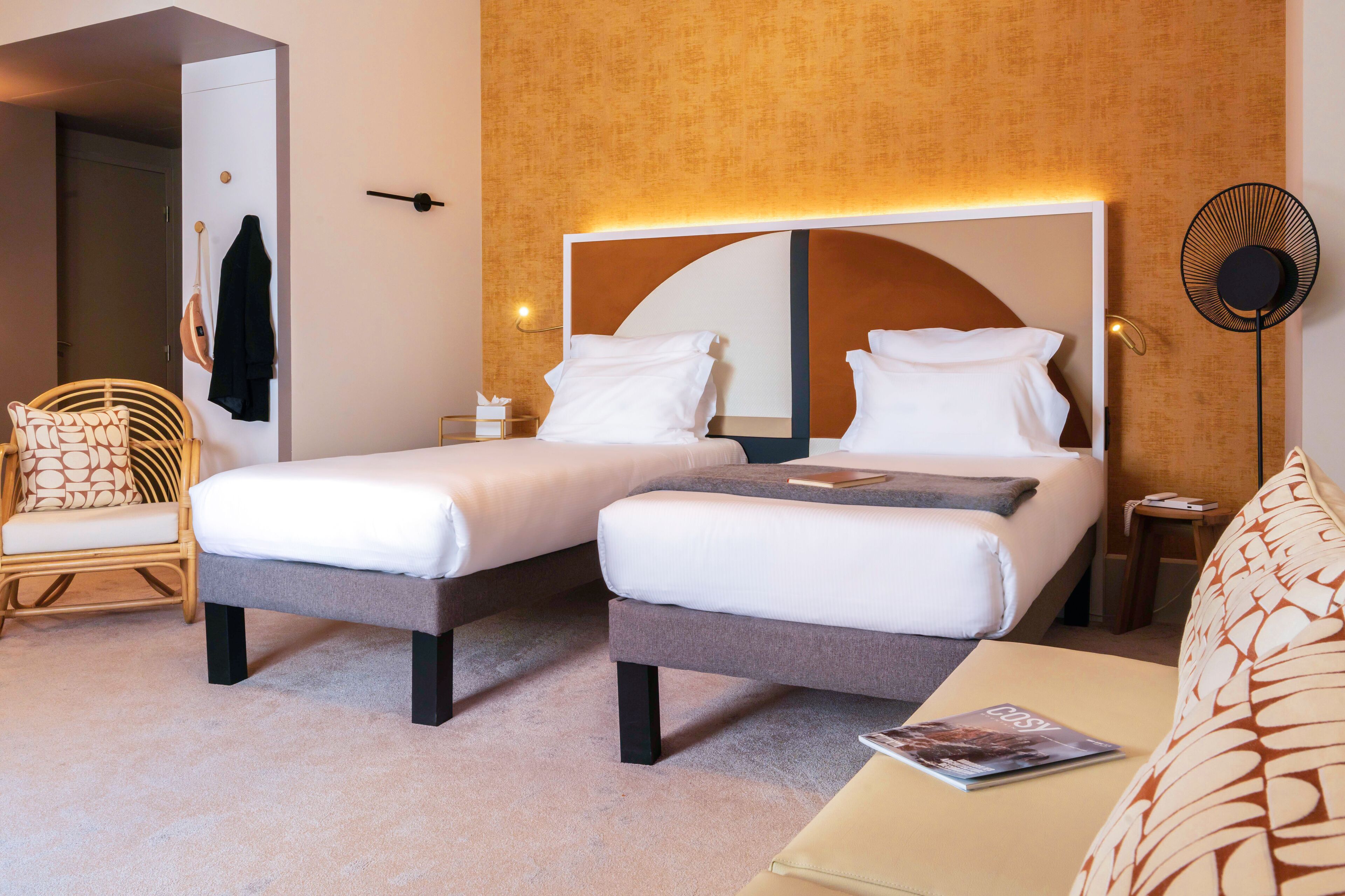 A hotel room with two beds, a sitting area, a chair, a bedside table, a wall-mounted fan, and a door leading to another room.
