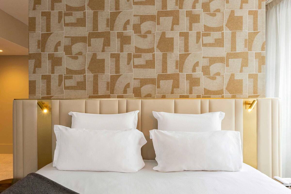 A hotel room with a large bed, a headboard with light-colored panels, and a geometric patterned wall above the bed.