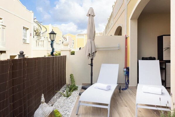 A small outdoor area with two white lounge chairs, a wooden fence, and a small garden with white pebbles and green plants. - Home2Book Palm Beach & Relax (Palm-Mar)
