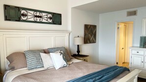 Un lit bien fait, recouvert d’une courtepointe matelassée, avec un plaid bleu et plusieurs oreillers. On y trouve une table de chevet avec une lampe, un élément décoratif fixé au mur, et une porte donnant sur une autre pièce.