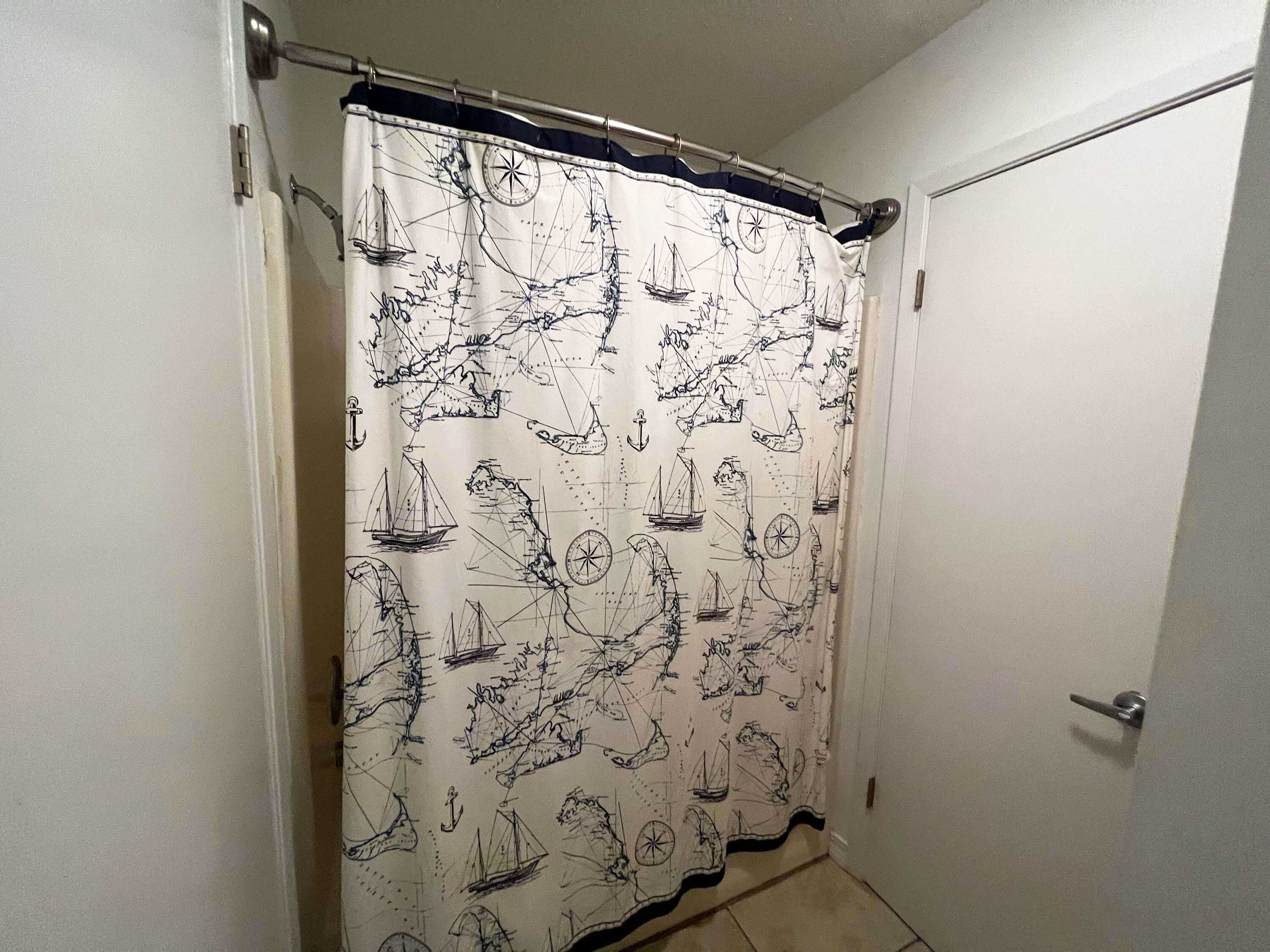 Un baño con puerta blanca, cortina de ducha blanca con motivos náuticos y suelo de baldosas.