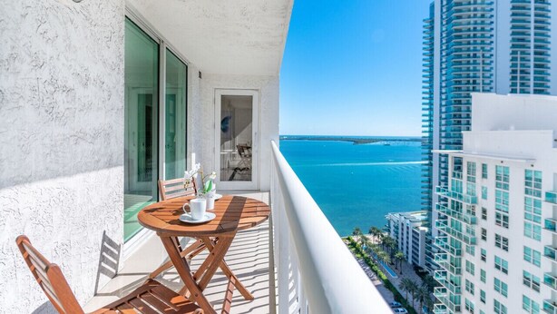 A balcony with a wooden table and chairs, overlooking a clear blue sea and a modern high-rise building.