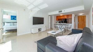 A modern living room with a sofa, a coffee table, and a TV. There is a kitchen area with wooden cabinets and a dining table.