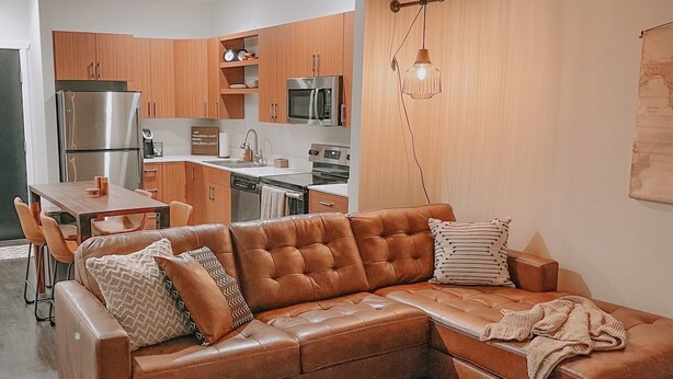 A modern living room with a brown leather sectional sofa, a dining table with chairs, and a kitchen area with wooden cabinets and stainless steel appliances. - Modern and Cozy Downtown TC Condo. Walk To Beaches, Bars, Restaurants & More. (Traverse City)