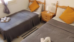 A room with two beds, each with a grey bedspread and a single yellow pillow. There is a nightstand with a lamp and a power outlet between the beds. A pair of towels is folded on the floor. - Apartment 450 metres from Playa del Cura (Torrevieja)