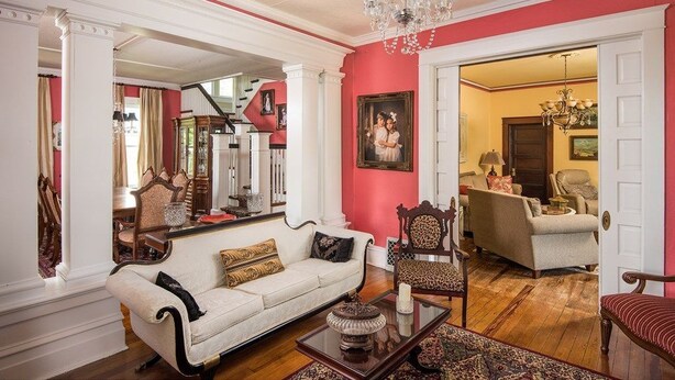 Un salón espacioso con un sofá blanco, un sillón estampado y una mesa de centro. Se ve una escalera al fondo que conduce a otra habitación.