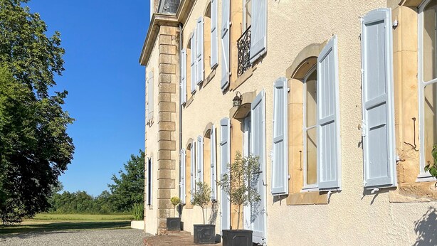 Un bâtiment doté de nombreuses fenêtres et de volets, un espace pavé agrémenté de plantes en pot et un canapé en osier avec des coussins.