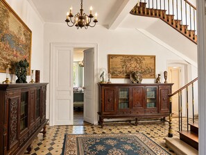 A grand hallway with a staircase, ornate wooden furniture, and a chandelier.