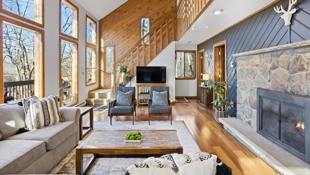 A spacious living room with a stone fireplace, wooden ceiling, and large windows.
