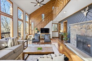 A spacious living room with a stone fireplace, wooden ceiling, and large windows. - Karaoke!•LUX French Chalet•HotTub•Pool Table•FirePit•Pets•AC•Beach+Lake Access! (Lake Harmony)
