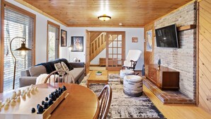 A cozy living room with a wooden ceiling, a fireplace, a TV mounted on the wall, and a chessboard on the table.