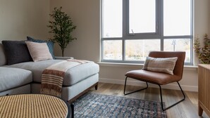 Een moderne woonkamer met een grijze bank, een bruine leren fauteuil en een groot raam met uitzicht.