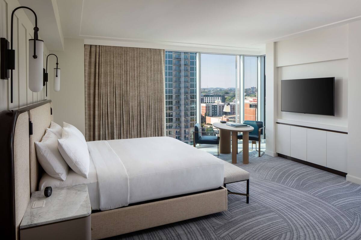 A hotel room with a large bed, a flat-screen TV, a small table, and a view of the city through large windows.