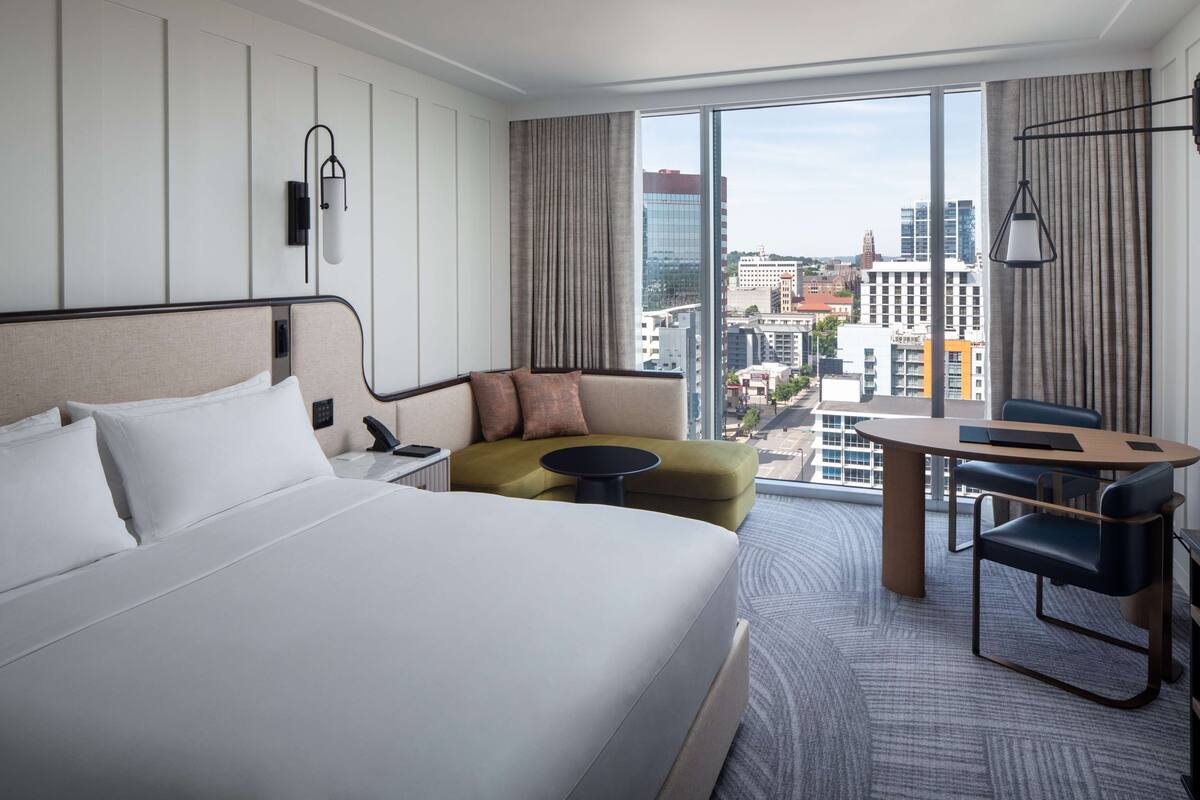 A hotel room with a large bed, a sofa, a small table, and a view of the cityscape.