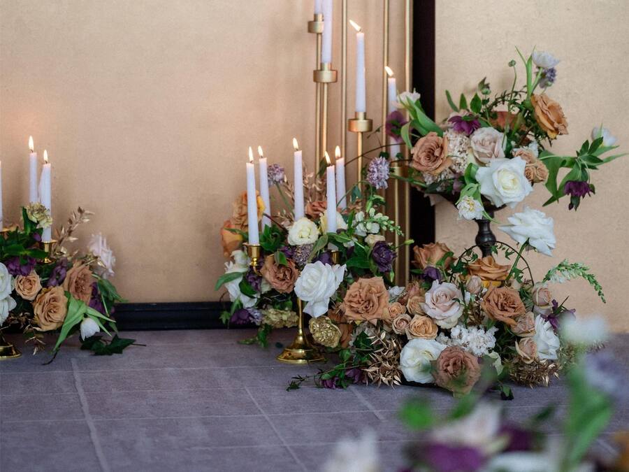 En række høje, hvide lys med gyldne fodstykker, omgivet af blomsterarrangementer.