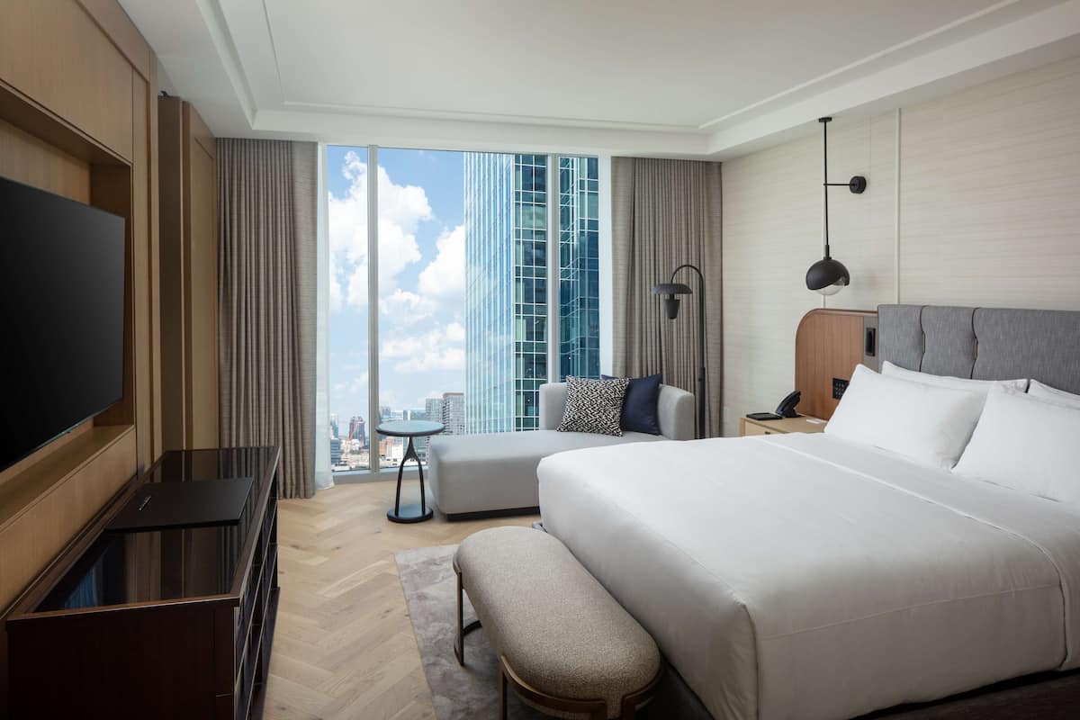 A modern hotel room with a large bed, a flat-screen TV, a sofa, and a city view.