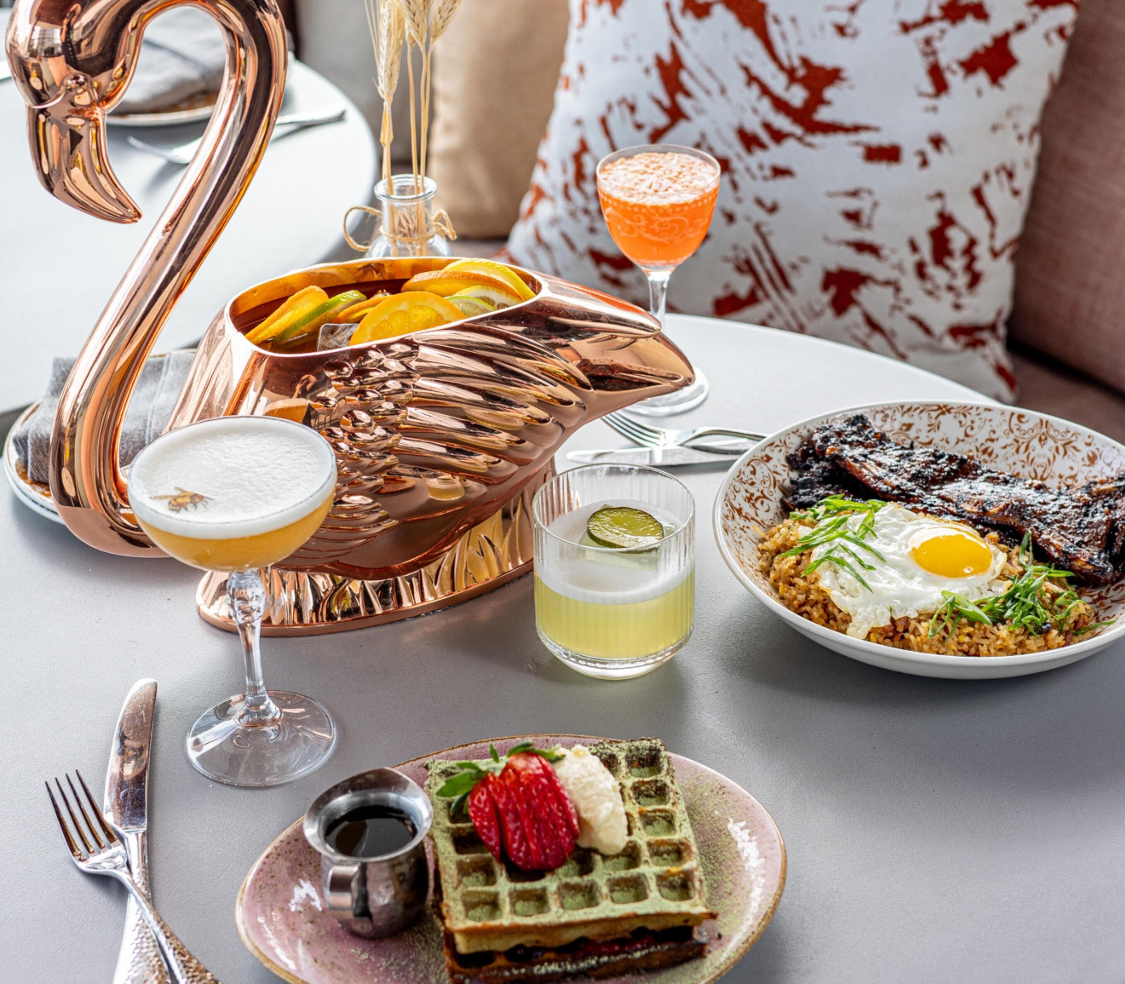 A dining table with a swan-shaped copper pitcher, a waffle with strawberries, a plate of fried rice with egg, a cocktail glass, and a small glass of beer.