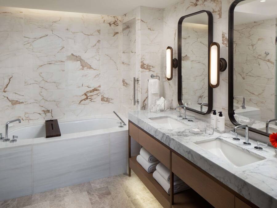 A modern bathroom with a marble wall, a bathtub, a vanity with two sinks, and a large mirror.