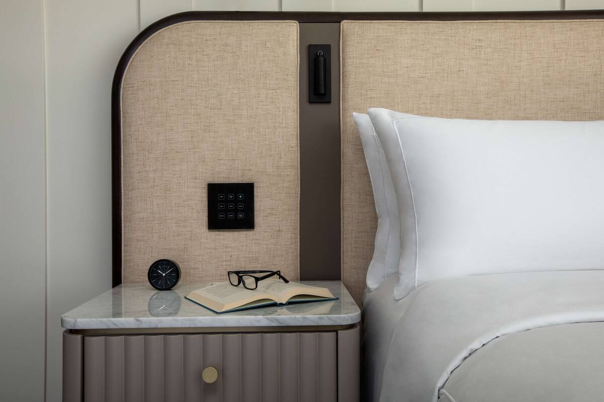 A neatly arranged hotel room with a bed, a bedside table with a clock, glasses, and an open book.