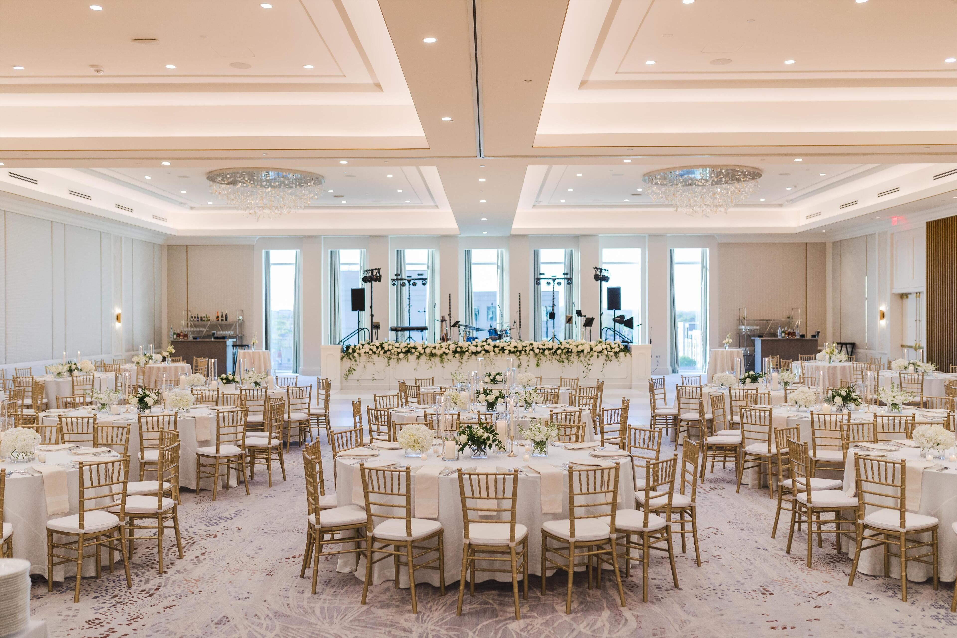 Ein Festsaal mit runden Tischen, goldenen Stühlen und floralen Tischdekorationen.