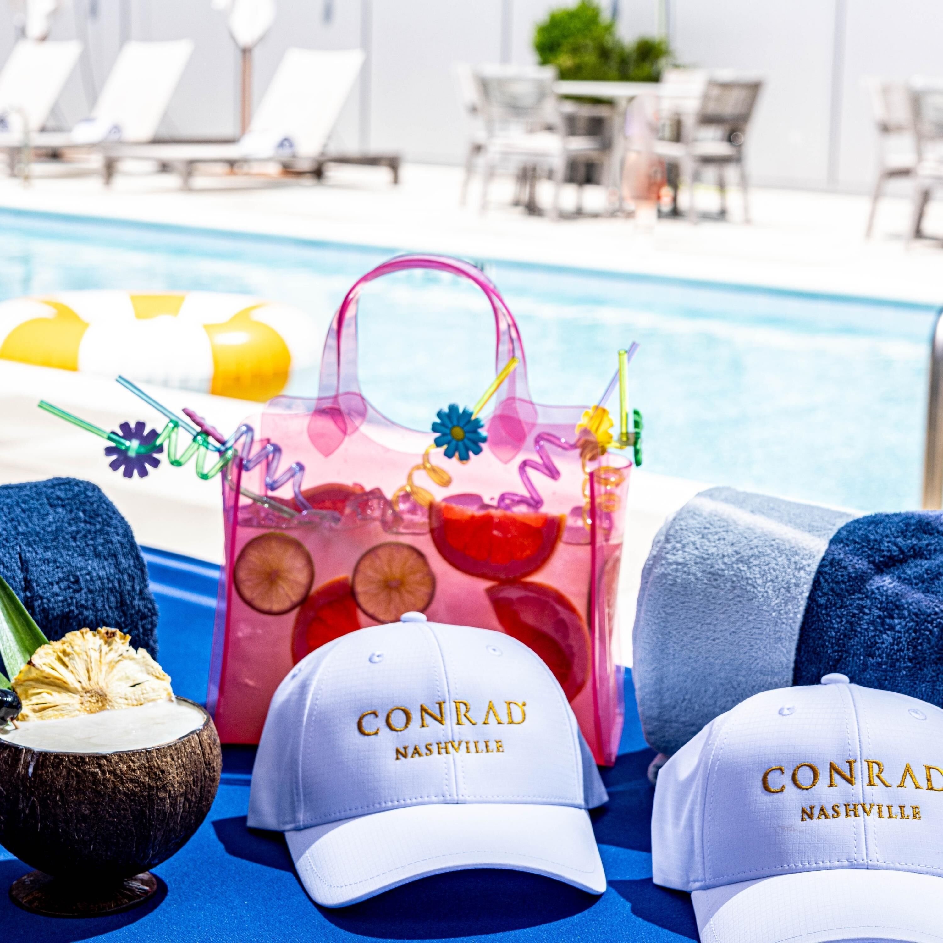 A poolside setting with lounge chairs, a drink with fruit slices, a tote bag, and two caps with "CONRAD NASHVILLE" text.