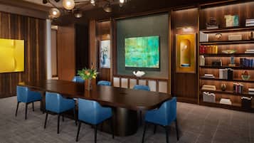 Uma sala de conferências com uma mesa comprida, cadeiras azuis, uma estante de livros e uma grande pintura abstrata.