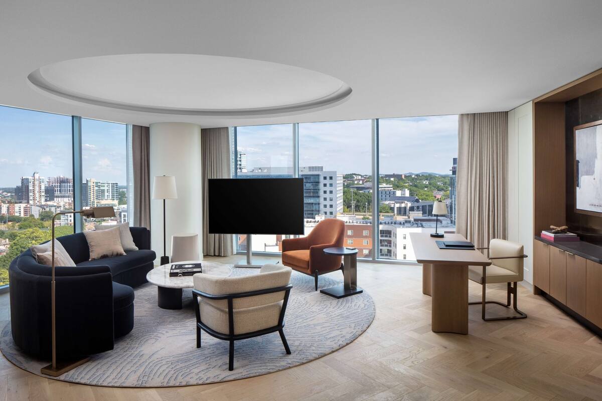 A modern hotel room with a large city view, a flat-screen TV, a sofa, a chair, a coffee table, and a desk.